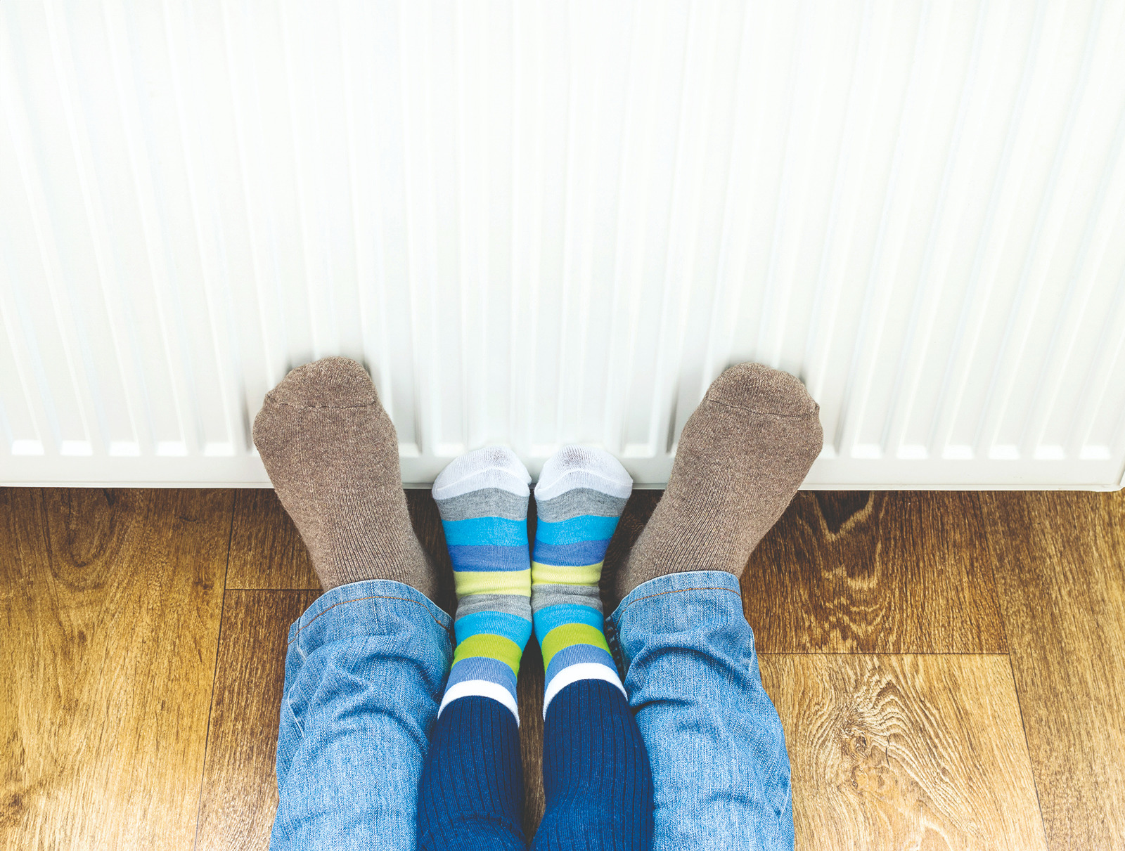 Zwei paar Füße an eine Heizung gelehnt, das größere Paar mit braunen Socken und einer Jean, das kleinere Paar mit bunten Ringelsocken.